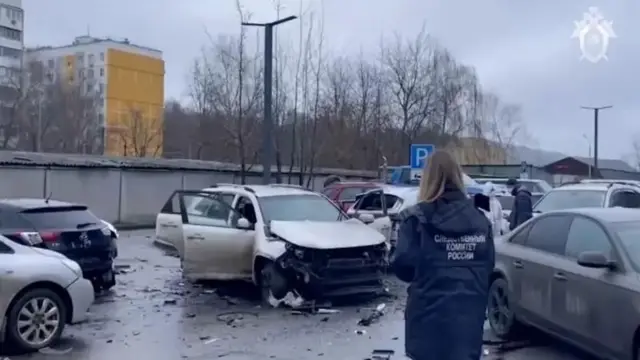 Фото підірваного авто на парковці, спиною до камери стоїть жінка у куртці з написом Слідчий комітет росії