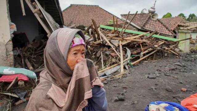 Seorang perempuan dengan latar bangunan yang hancur sedang meratapi dampak dari erupsi Gunung Semeru.