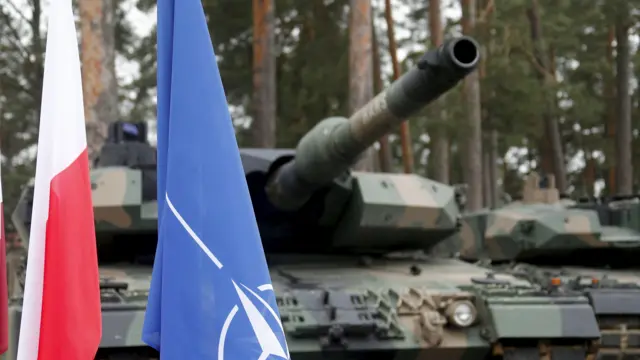 Dwa czołgi zaparkowane są za flagami NATO i Polski, w tle widać sosny.