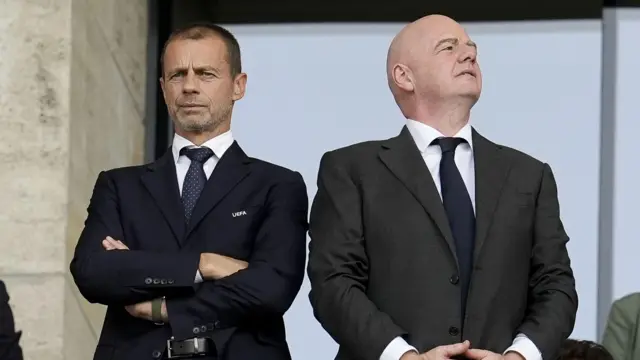 Przewodniczący UEFA Aleksander Čeferin (po lewej) i Gianni Infantino. Obaj są ubrani w garnitury. Mężczyźni nie patrzą na siebie; Čeferin krzyżuje przed sobą ramiona.