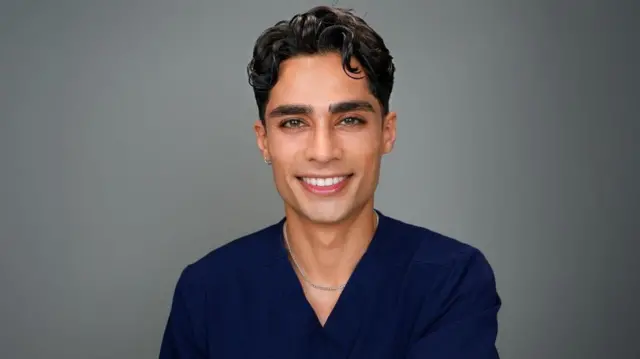 El Dr. Zayn sonríe a la cámara. Lleva un uniforme quirúrgico azul y una cadena de plata alrededor del cuello. Tiene dientes blancos, piel aceitunada y un ligero rizo en su cabello castaño. También lleva un pequeño aro de plata en la oreja.