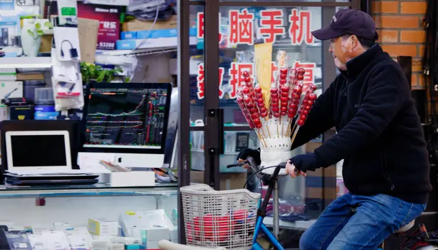 Людина сидить на велосипеді з кошиком, а на кермі закріплений великий букет із цукерок на паличках. На задньому шо плані – вітрина магазину з комп’ютерною технікою: ноутбуки, монітори, коробки з електронікою. Один із моніторів показує графік біржових котирувань. На скляних дверях магазину видно червоні китайські ієрогліфи.
