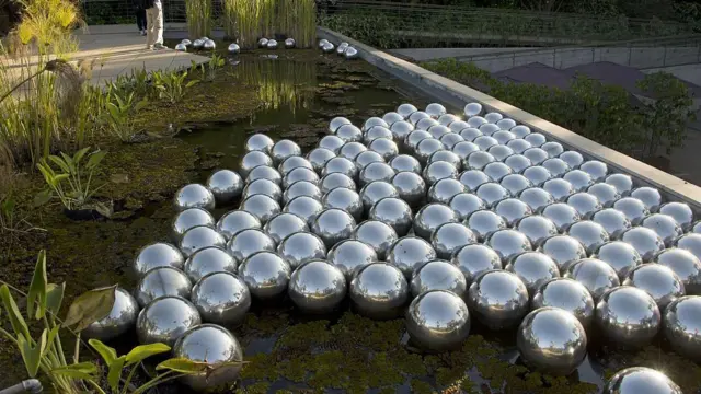 Instalação com dezenas de esferas espelhadas prateadas sobre a superfície de um lago, cercadas por vegetação e passarelas.