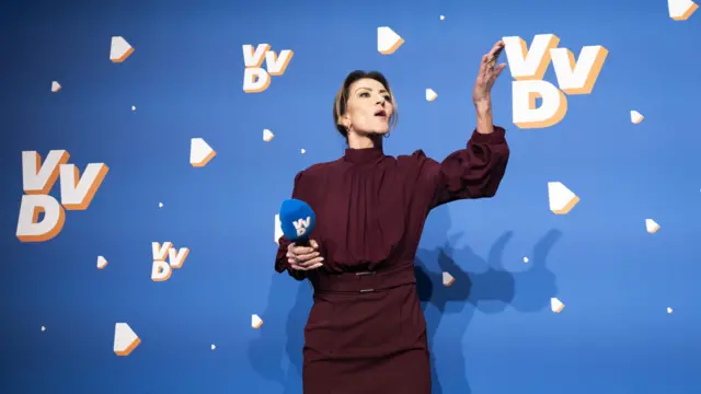 Дилан Йешильгёз на фоне символики VVD с бордовом платье радостно стоит с микрофоном в руке.