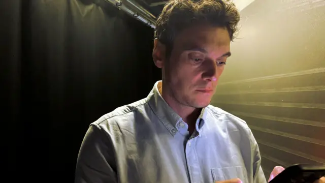 Joe Tidy mirando un móvil. Tiene el pelo castaño corto y una camisa azul claro. 
