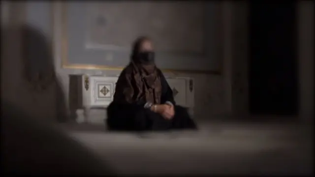 بنفشه چهارزانو روی زمین نشسته و دستانش را در هم قفل کرده است. لباسهای تیره به تن دارد؛ چادرش بنفش تیره و مانتویش سیاه است و ماسک سیاه به صورت زده است