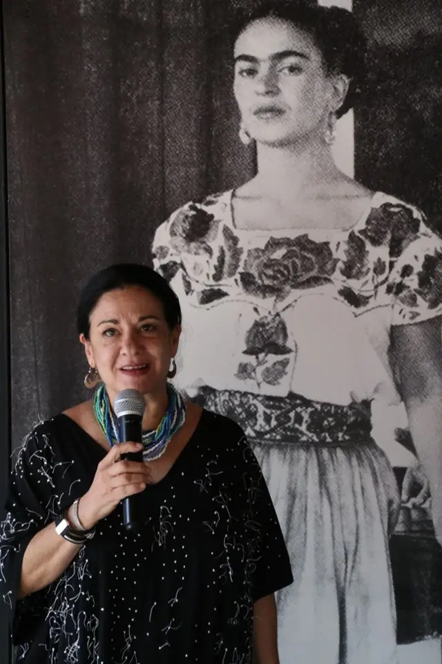 Cristina Kahlo Alcalá lleva un vestido negro con puntos blancos, aretes y un collar de varias vueltas azul y blanco. En su mano tiene un micrófono y detrás de ella hay una foto de Frida Kahlo, quien mira la cámara. Está vestida con la ropa tradicional de Tehuana
