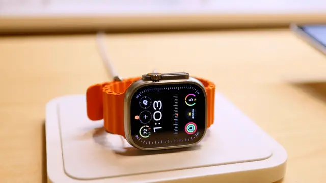 Apple Watch com mostrador preto e pulseira laranja sobre uma mesa, marcando 1h03