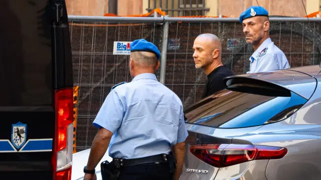 Mężczyznę w czarnym T-shircie wyprowadza z srebrnego radiowozu dwóch policjantów ubranych w jasnoniebieskie mundury.