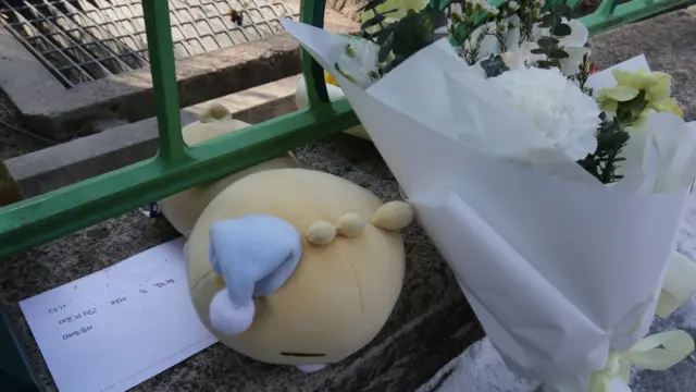 A school wall with a card that says, "Don't be sick, baby. Close your eyes comfortably. I'm sorry," a doll, and a bouquet of flowers.