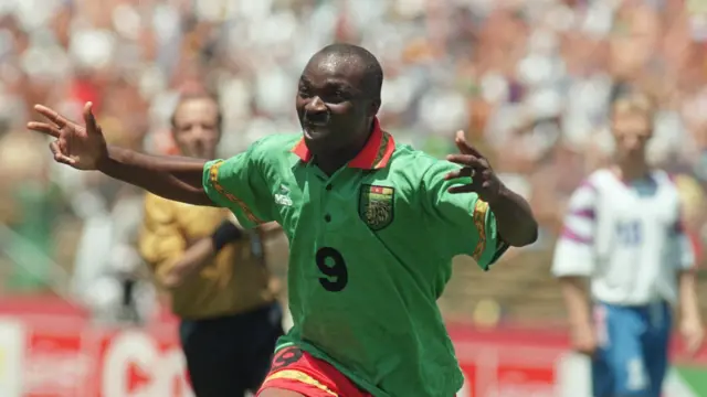 Roger Milla court alors qu'il célèbre le fait d'avoir marqué contre la Russie lors de la Coupe du monde 1994 en écartant les bras et en arborant un large sourire aux lèvres. Milla porte un maillot camerounais vert avec un col rouge et un chiffre neuf en gras sur la poitrine, avec un short rouge. À l'arrière-plan, on peut voir un arbitre vêtu d'un maillot jaune et d'un short noir et un joueur russe vêtu d'un maillot blanc et d'un short bleu devant une foule.