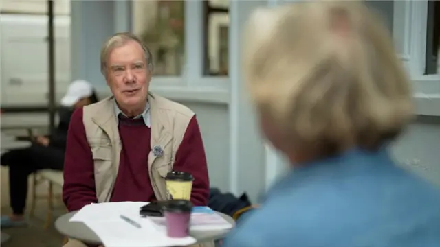 گوردون با جاکت سرخ در کافه مقابل زنی با موهای طلایی رنگ نشسته است. زن پشت به کمره است. 