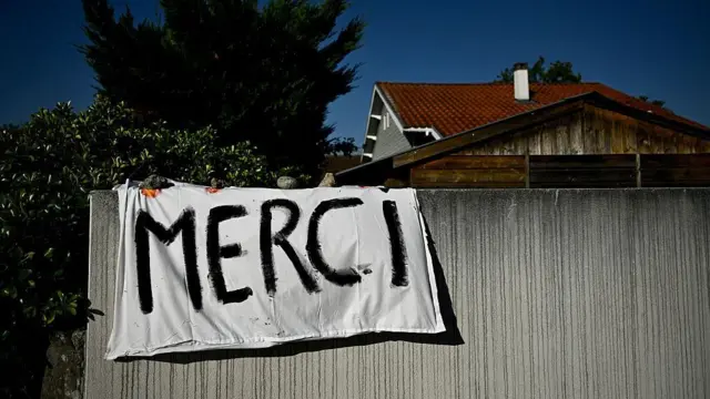 一軒家の塀に黒いスプレーで「MERCI（ありがとう）」と書かれた白いシーツがかけられている