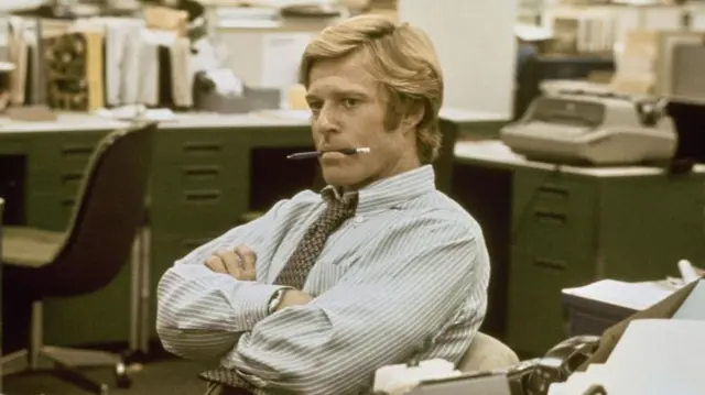 Amerykański aktor Robert Redford w niebiesko-białej koszuli w paski z wzorzystym krawatem, z założonymi rękami i ołówkiem w ustach, w rekonstrukcji biur „The Washington Post” w filmie „Wszyscy ludzie prezydenta”, nakręconym w Burbank Studios w Burbank w Kalifornii w 1976 roku.