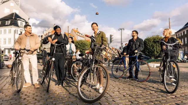 Un grup de turiști bucuroși împreună cu ghida lor stau fiecare lângă o bicicletă pe o stradă cu piatră cubică.