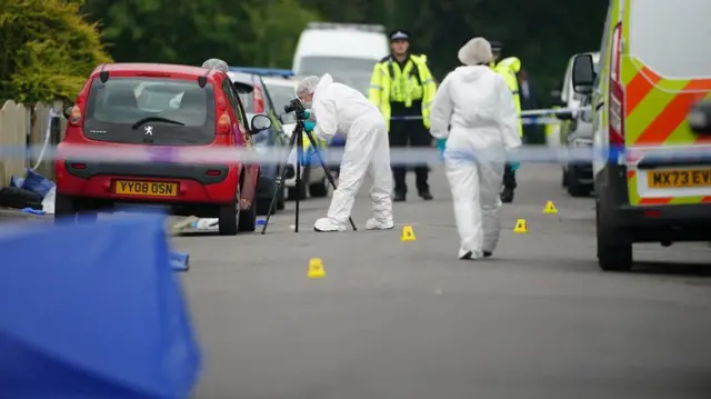 A forensic officer in white protective gear photographs the road surface, with two uniformed constables looking on 