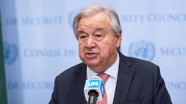 Katibu Mkuu wa Umoja wa Mataifa Antonio Guterres akizungumza kwenye kipaza sauti chenye nembo ya Umoja wa Mataifa. Amevaa suti nyeusi yenye tai nyekundu yenye madoadoa. Nyuma kuna bango lenye ukungu lenye herufi zinazoonekana kidogo zinazoonyesha baraza la usalama.