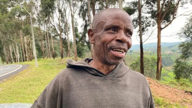 Umugabo uhagaze urimo kuvuga, inyuma ye hari ibiti mu ishyamba