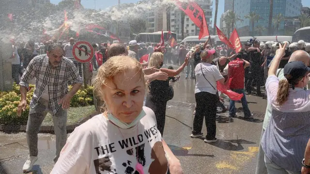 Önde bir kadın saçları ıslanmış durumda. Arkada çok sayıda kişi polis barikatının önünde duruyor.