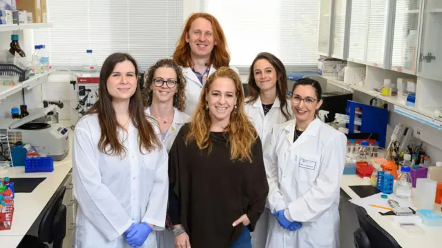 Grupo de seis cientistas — cinco mulheres e um homem — posando para uma foto de grupo, a maioria de jaleco branco em um ambiente de laboratório