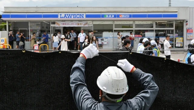 Pracownik w kombinezonie i hełmie ochronnym rozwiesza gęstą, ciemną siatkę zasłaniającą widok. U góry zdjęcia, za siatką, widzimy grupę osób w swobodnych strojach przed przydrożnym supermarketem.