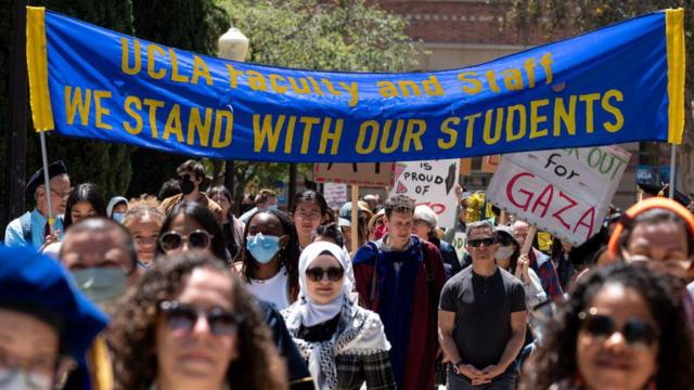 Alguns docentes e funcionários da  UCLA marcham sob uma faixa que diz 'Estamos com nossos estudantes' no campus universitário de Los Angeles na segunda-feira, 29 de abril de 2024