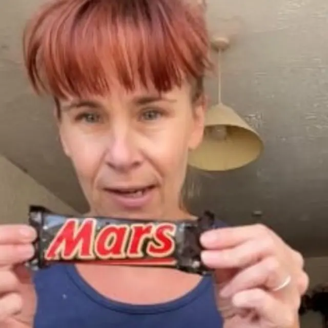 Жінка тримає в руках старий батончик Mars. Вона одягнена в темно-синю майку; у неї руде волосся, зібране догори, і густий чубчик.