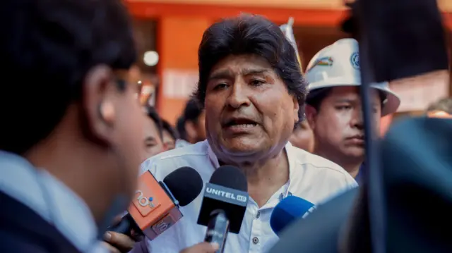 Evo Morales respondiendo a la prensa, tiene dos micrófonos delante. Detrás hay un hombre con casco de obra y otras personas a las que apenas se ve.
