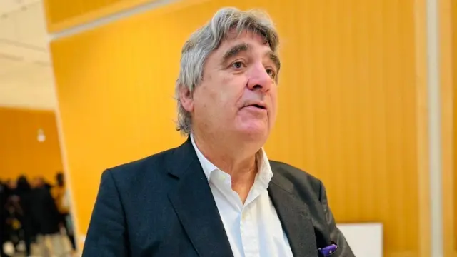 Bernard Maingain arimo kuvuga, yambaye ikoti ry'ubururu bwijimye bujya kuba umukara, n'ishati y'umweru