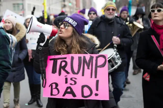 A marcher with a hat that says "Forward" and a megaphone in a group holds a sign that says "Trump is BAD!" as she walks in front of Trump International Tower during the Woman's March