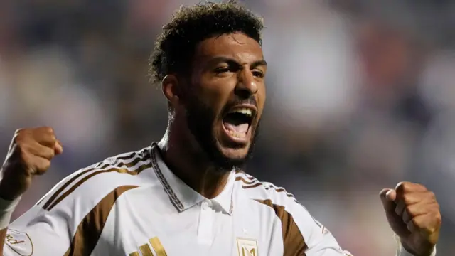 Denis Bouanga, vêtu d'un maillot blanc et or du LA FC, lève les deux poings en signe de triomphe et crie en célébrant un but.