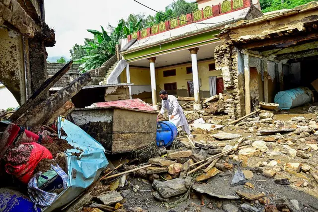বন্যার পানির তোড়ে ভেঙে যাওয়া একটি বাড়ির সামনে মালপত্র সরানোর কাজ করছেন এক ব্যক্তি 