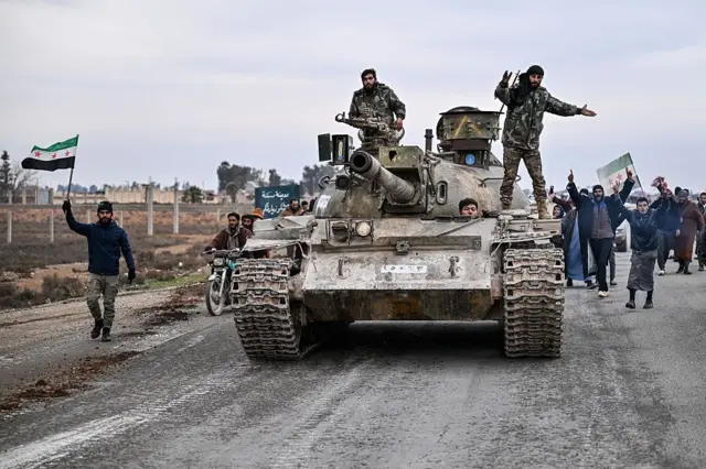 دبابة عليها عسكريون سوريون، ووراءهم عدد من الأشخاص يحملون علم سوريا ذو النجوم الثلاث الحمراء.