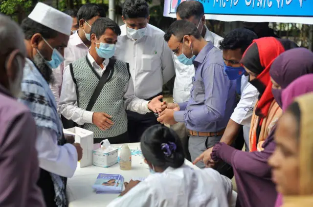 এক ব্যক্তির আঙুল থেকে রক্ত নিয়ে ডায়াবেটিস পরীক্ষা করছেন একজন। পাশে দাঁড়িয়ে আছেন অনেকে এবং সবারই মুখে মাস্ক। টেবিলের ওপর রক্ত পরীক্ষার কিট রাখা।