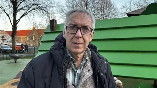 La fotografía muestra a un hombre de cabello gris, con gafas y un abrigo con capucha negro, parado frente a un parque de juegos infantiles en un parque.