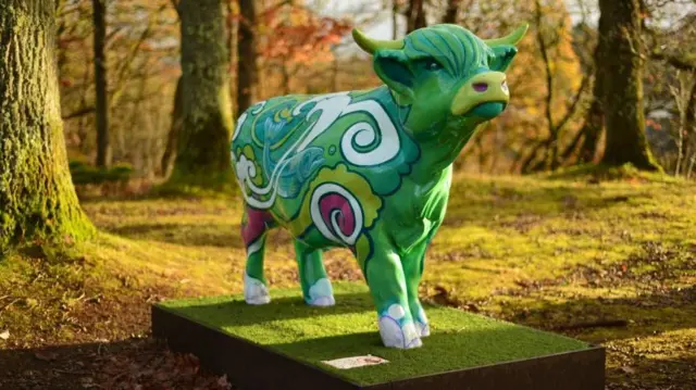 Una escultura de una vaca de las Highlands, pintada con colores brillantes, sobre una plataforma rectangular cubierta de césped artificial. La vaca está decorada en tonos verdes intensos con patrones en espiral en blanco, azul, rosa y amarillo, lo que le da un aspecto psicodélico y artístico.