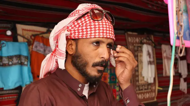A man applying black eyeliner wearing a brown shirt and a traditional scarf or Arabic man's headdress tied around his head which is red and white.