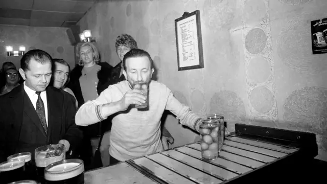 Un hombre en un bar, rodeado de personas, se toma un vaso lleno de huevos.