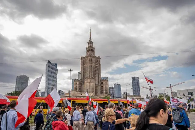 Учасники маршу з польськими прапорами зібралися в центрі Варшави. На тлі видно Палац культури і науки та сучасні хмарочоси. Частина учасників тримає банери, а над натовпом майорять десятки біло-червоних прапорів. Небо затягнуте хмарами, що надає сцені похмурого вигляду.