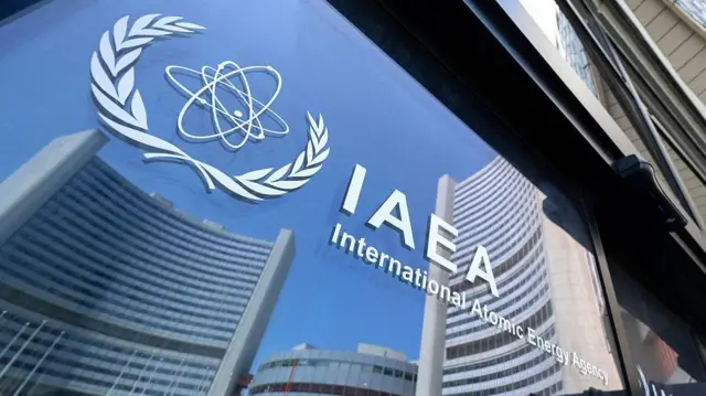 На стеклянной двери рядом с символом агентства белым цветом выгравирована аббревиатура IAEA (МАГАТЭ). В отражении стекла видны окружающие здания с изогнутыми фасадами и голубое небо.