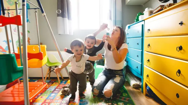 Mãe brincando de bolinha de sabão com os dois filhos. Ela está ajoelhada no chão e os filhos sorriem e agitam os braços, num quarto infantil bem colorido