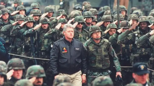 Білл Клінтон у чорній куртці та світлих штанах з американськими миротворцями у Тузлі, Боснія та Герцеговина