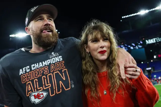 Taylor Swift e Travis Kelce em um jogo de futebol americano. O braço dele está sobre o ombro dela e ela segura o dedo médio dele com a mão esquerda.