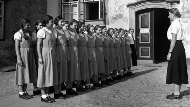 Meninas uniformizadas enfileiradas em frente a uma mulher em foto em preto e branco