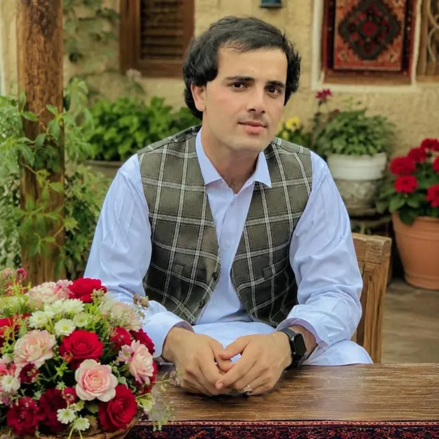 اسلم سایل یوه کلاسيک بیګراونډ کې ناست دی، کیلي دار واسکټ یې اغوستی دی.