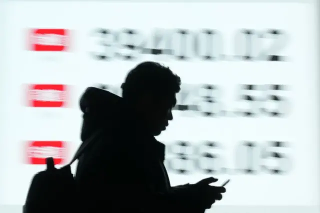 La silueta de una persona viendo un teléfono pasa frente a una iluminada valla que reporta la caída de los índices de la bolsa de valores de Tokio, con símbolos rojos seguidos de números. 