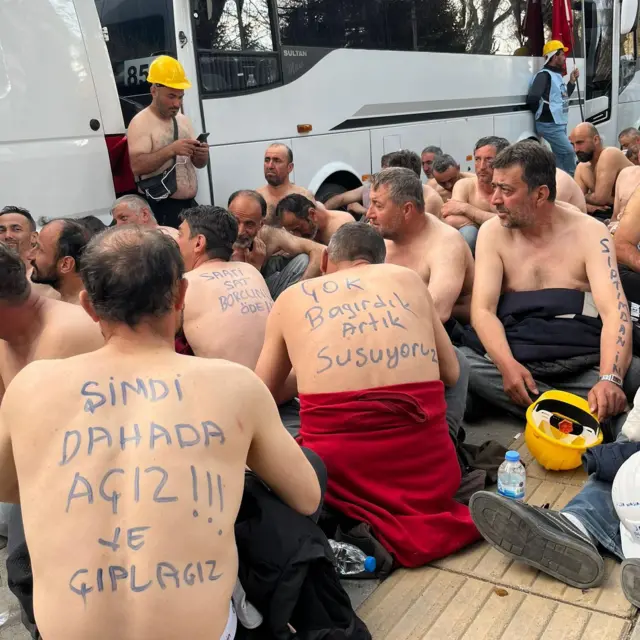 Üstü çıplak erkekler yerde oturuyor. Sırtalarında "Şimdi daha da açız ve çıplağız", "Çok bağırdık artık suruyoruz" gibi sloganlar yazılı. 