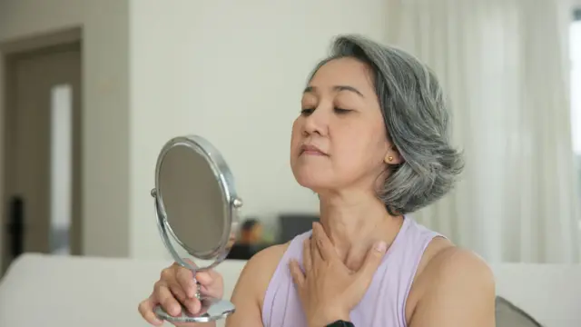 Una mujer de mediana edad se mira en un espejo de mesa las arrugas que se le empiezan a notar en el cuello. 