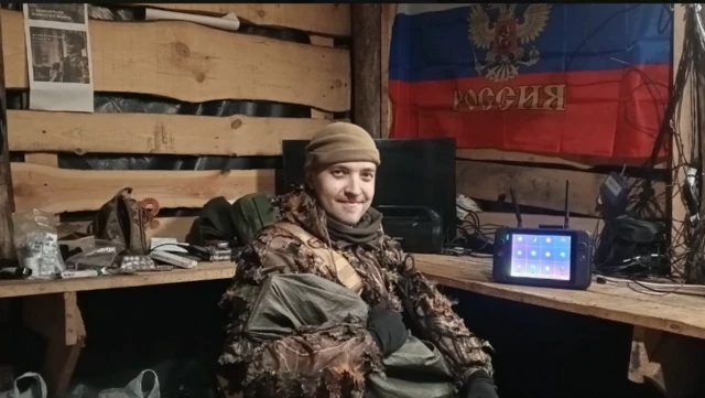 Человек сидит в блиндаже и улыбается на фоне флага России на стене