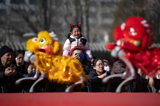 Люди смотрят на выступление четвероногих роботов, одетых в новогодние костюмы. В центре кадра девочка на плечах у мужчины.